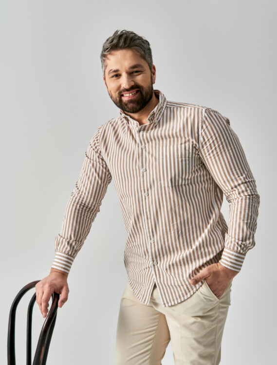 A stylish man with a beard standing confidently, holding a suitcase, and smiling with joy on a grey studio background.